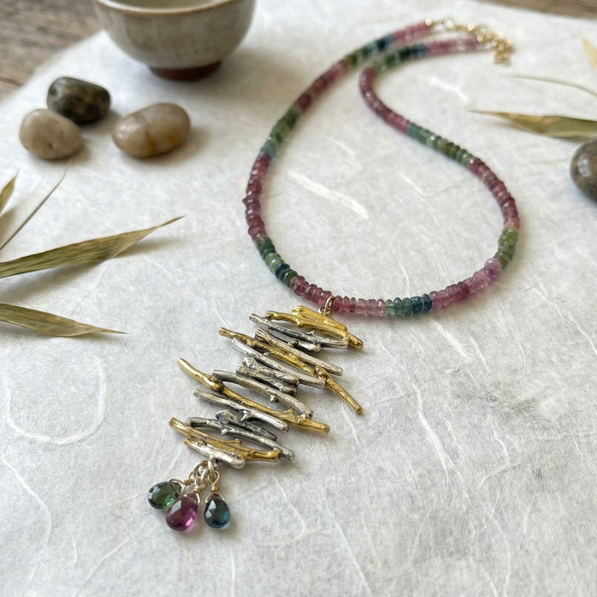 Necklace with multicolored beads and gold pendants on a textured white surface with stones and leaves.