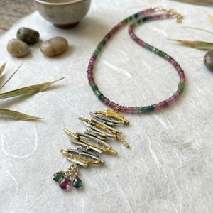 Necklace with multicolored beads and gold pendants on a textured white surface with stones and leaves.